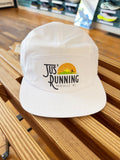 A white 5-panel cap with a 'Jus' Running Asheville NC' graphic on the front, resting on a wooden slatted bench in a retail environment.