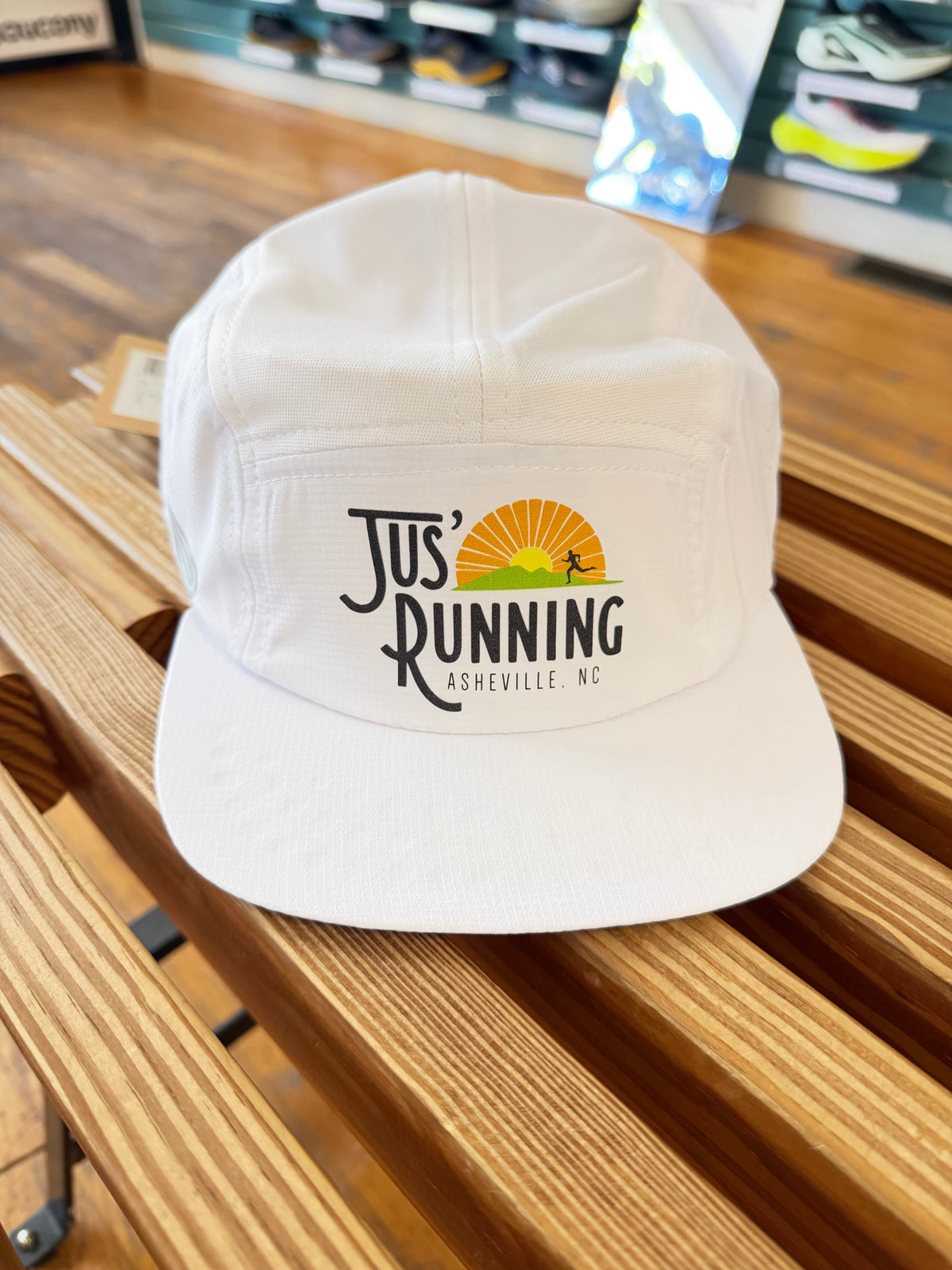 A white 5-panel cap with a 'Jus' Running Asheville NC' graphic on the front, resting on a wooden slatted bench in a retail environment.