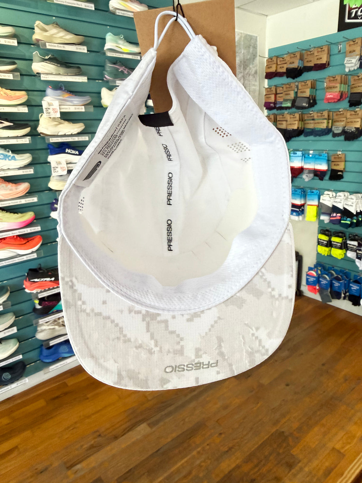 A white running cap with a subtle gray digital camouflage pattern on the brim, hanging on a display stand inside a shoe store. The brand name 'PRESSIO' is visible on the brim and inner label.