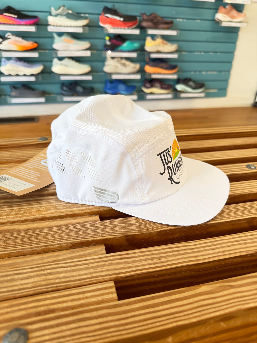 A white mesh-back running cap with a flat brim, featuring a 'Jus' Running' logo on the side, resting on a wooden slatted bench in a shoe store with a teal shoe display wall in the background.