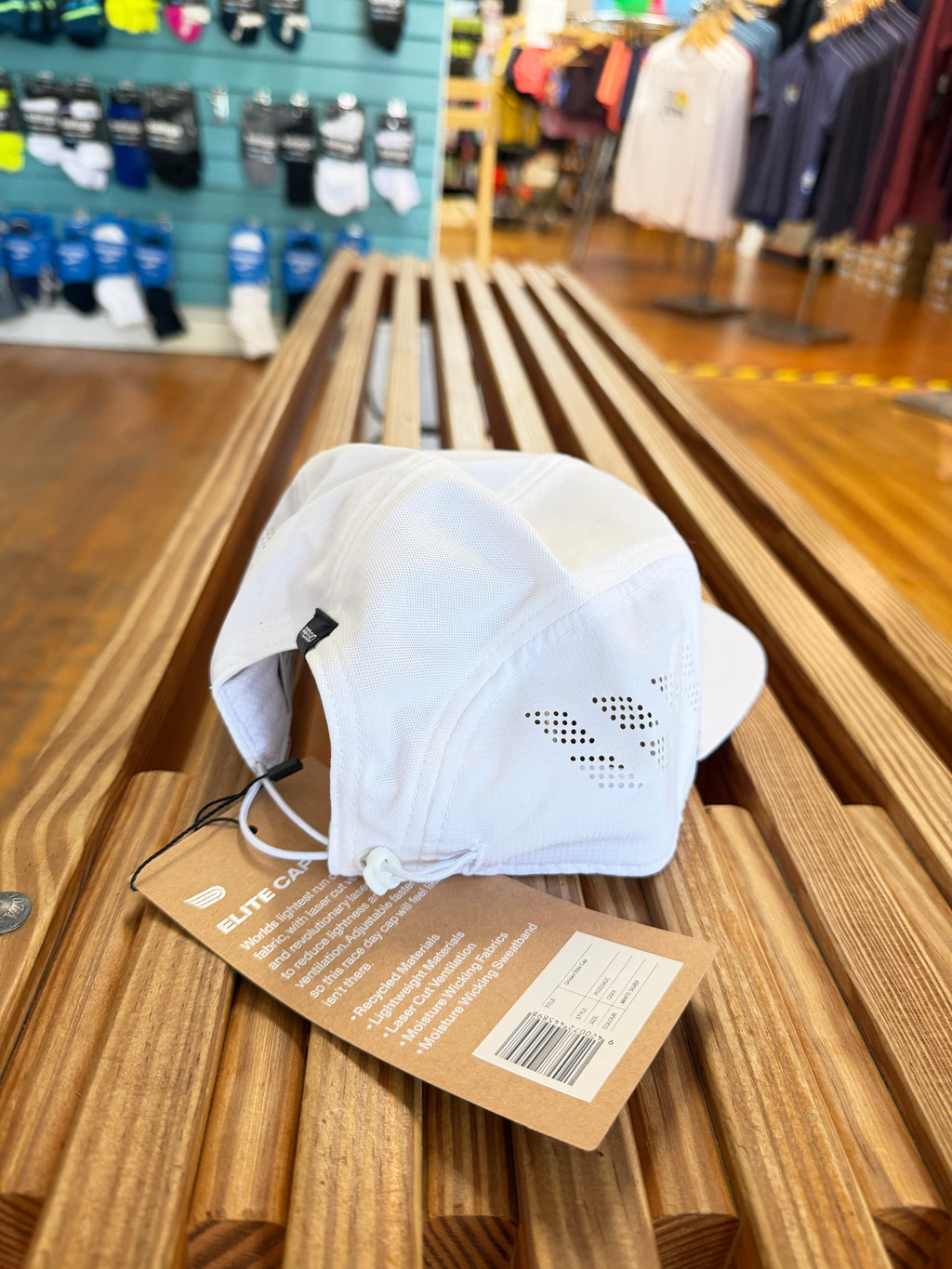 A white athletic cap with ventilation on the side, resting on a wooden slatted bench inside a retail store. A brown paper product tag is attached to the brim.