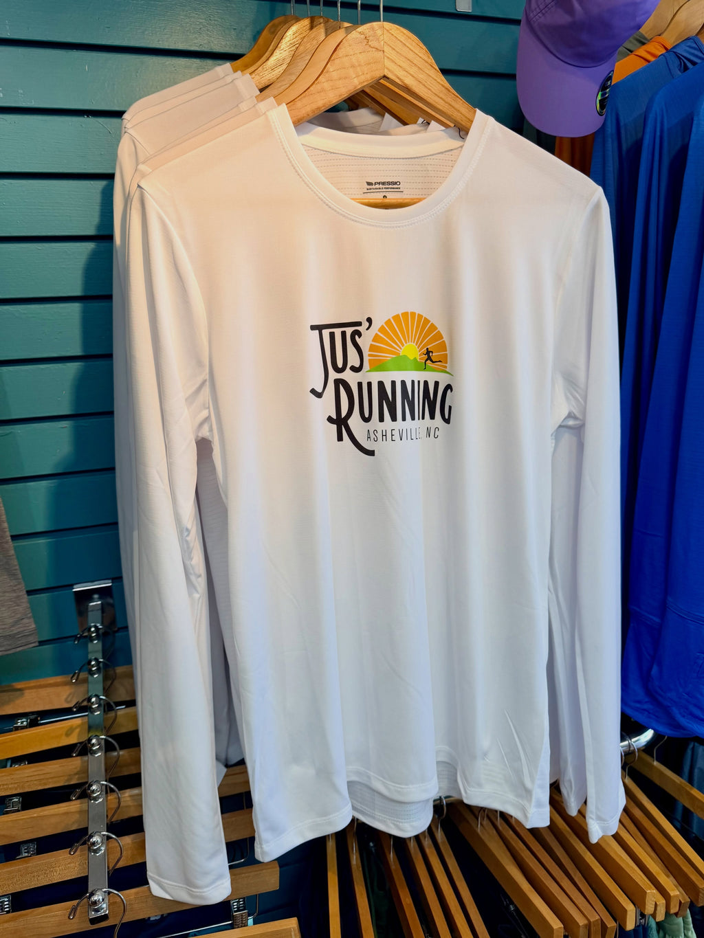 A white long-sleeve athletic t-shirt hanging on a wooden hanger against a teal slatwall. The shirt features a 'Jus' Running Asheville, NC' graphic on the chest with a black logo and a yellow/orange sunburst illustration.