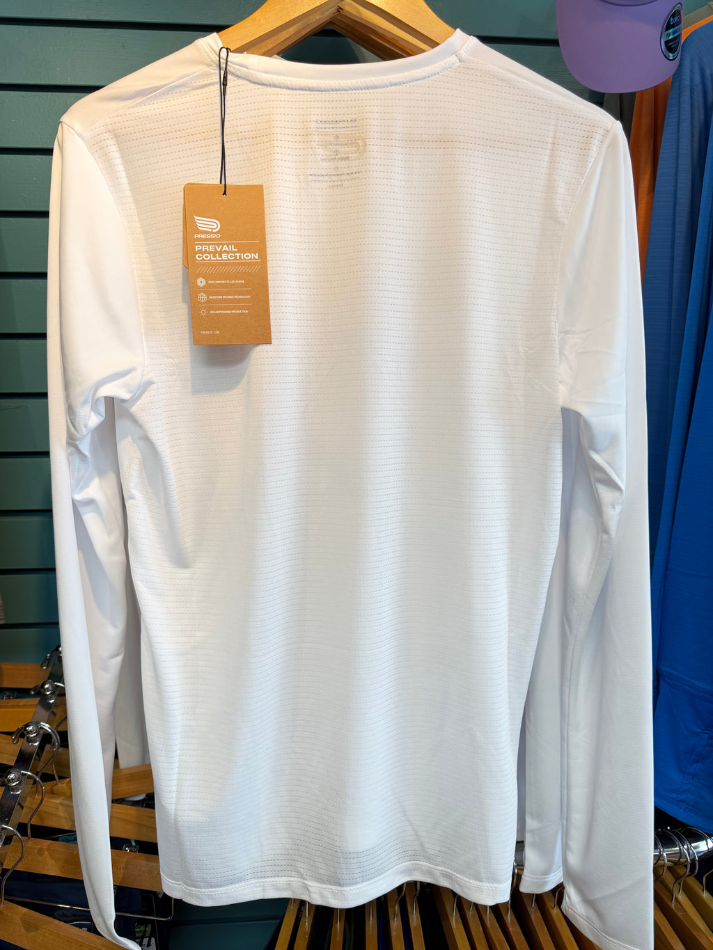 A white long-sleeve athletic shirt hanging on a wooden hanger, showing the back of the garment with a brown hangtag attached. The shirt has a subtle textured waffle pattern and is photographed against a dark teal slatwall background.