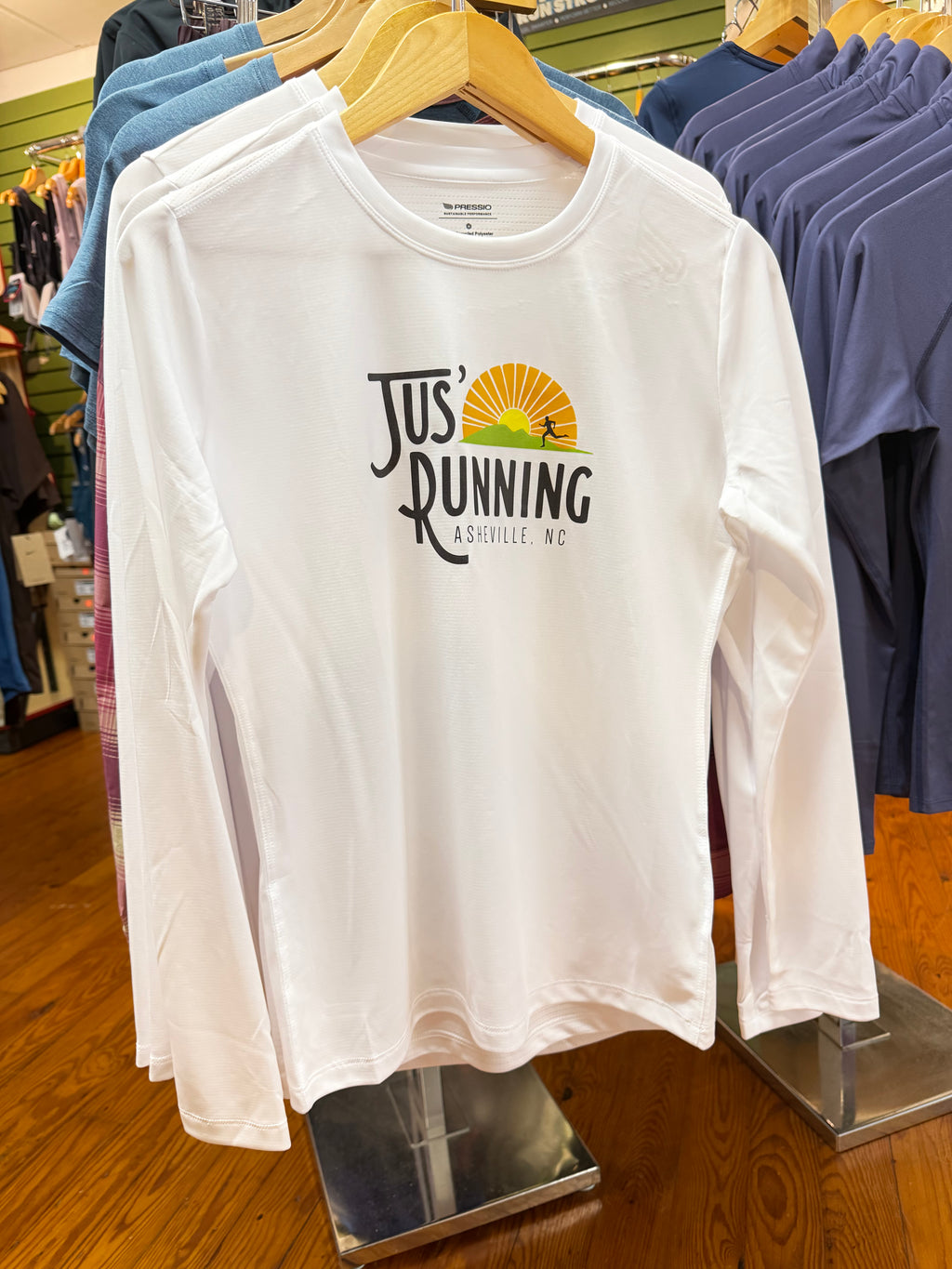 A white long-sleeve t-shirt hanging on a wooden hanger in a retail store, featuring a 'Jus' Running' graphic on the chest with a yellow and orange sun and green landscape.