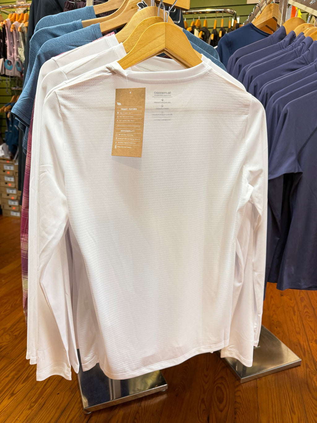 Back of white long-sleeve top hanging on a wooden hanger in a retail store, with a brown paper tag attached and other garments visible in the background.